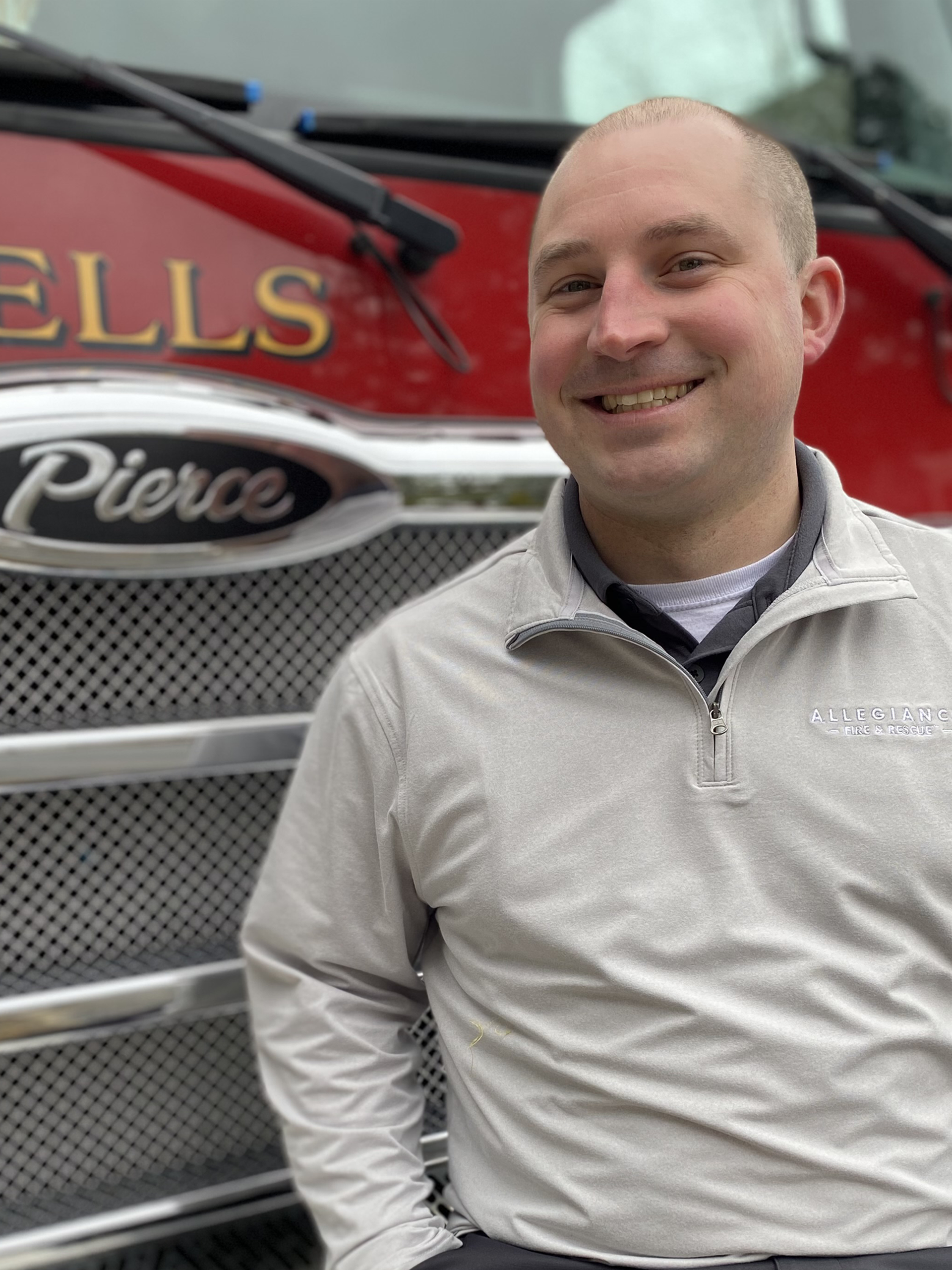 Bill Kimball, a professional Sales Rep sitting on the front bumper of the Wells, Pierce fire truck. He is wearing an off-white allegiance fire & rescue quarter zip shirt, clean shaven head and face, with a friendly smile.